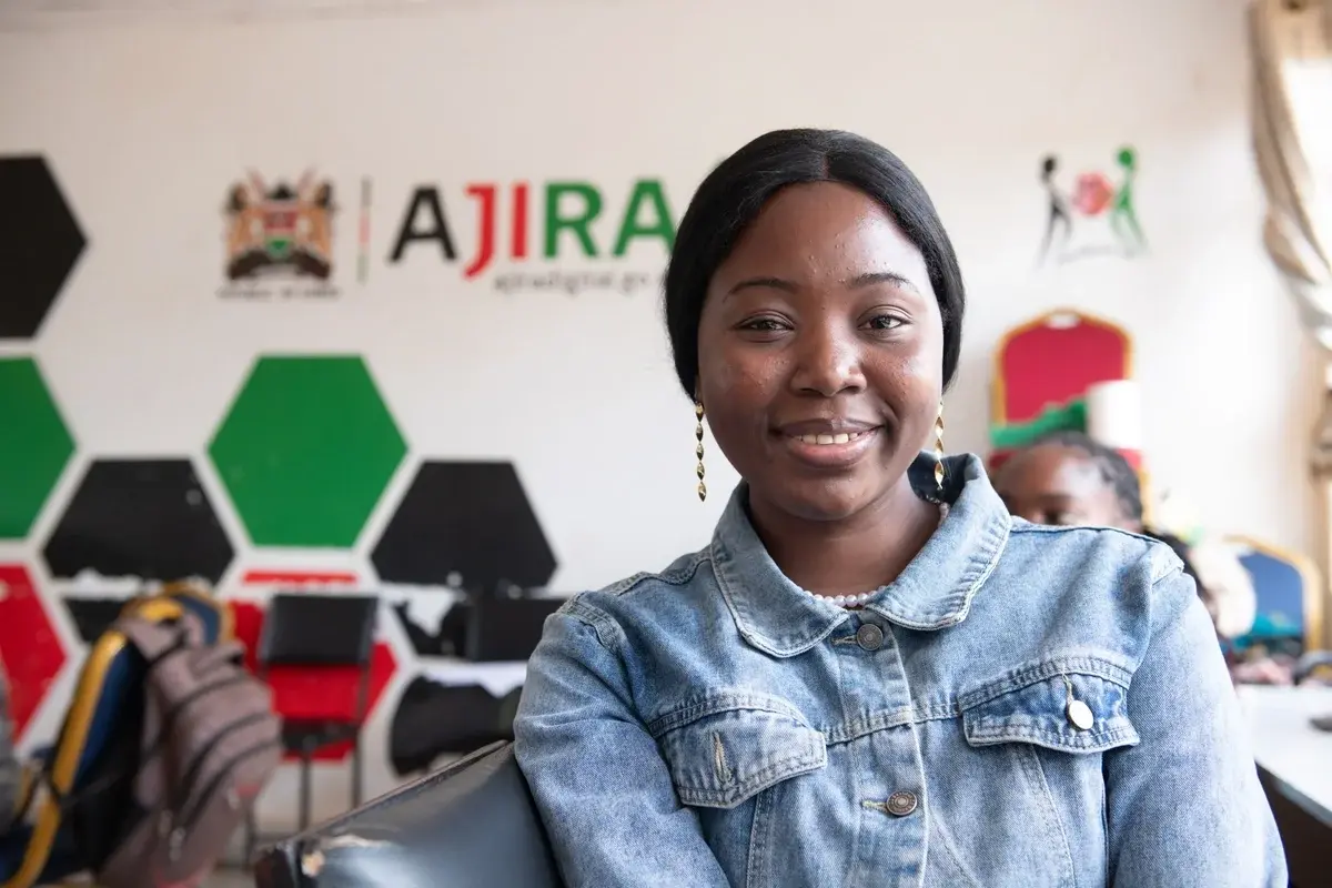 A young woman standing inside the Ajira office in Kenya, wearing a light blue button up jean jacket and smiling.